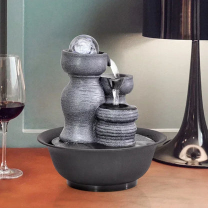 Small gray stone-like water fountain on a wooden surface with a glass of red wine and a lamp in the background.