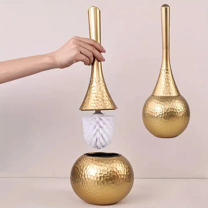 Gold toilet brush holder with a hand holding the handle against a light pink background