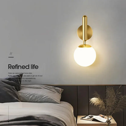 Modern wall lamp with gold finish and white glass shade in a bedroom setting.