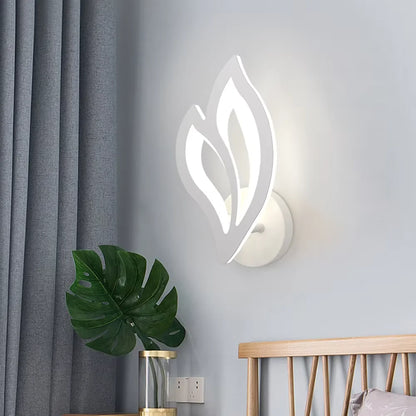 Modern wall sconce with leaf design on a light gray wall, next to a plant and wooden bed.